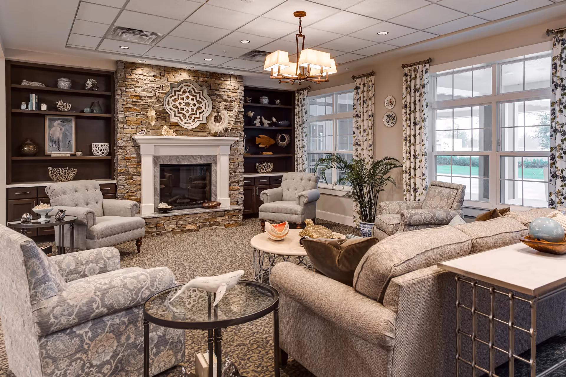 Cozy communal living room with upholstered chairs and sofas arranged around a stone fireplace and large windows.