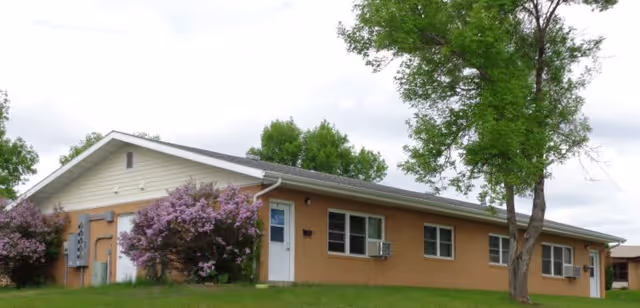 Single-story building with beige walls and a white door, surrounded by green grass, trees, and purple flowering bushes under a cloudy sky.