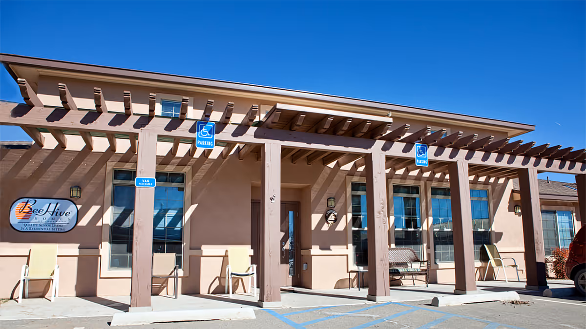 Exterior view of BeeHive Homes of Taylor Ranch showing the front entrance with a pergola structure, several chairs placed outside, handicap parking signs, and a clear blue sky.