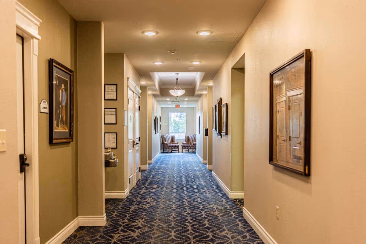 A well-lit hallway in an assisted living facility with beige walls and a patterned blue carpet. The hallway features framed artwork on the walls, recessed ceiling lights, and a water fountain on the left side. At the end of the hallway, two upholstered chairs are positioned near a window letting in natural light.