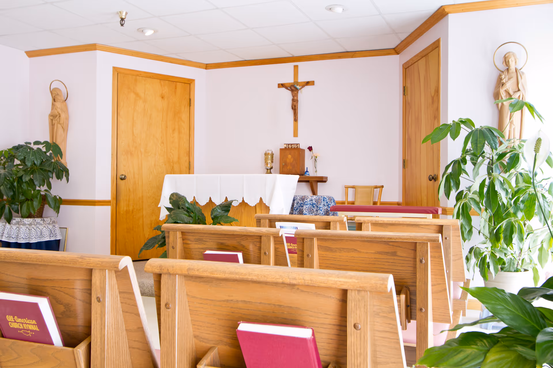 Small chapel-like interior with wooden pews facing an altar topped by a crucifix, flanked by statues and potted plants.