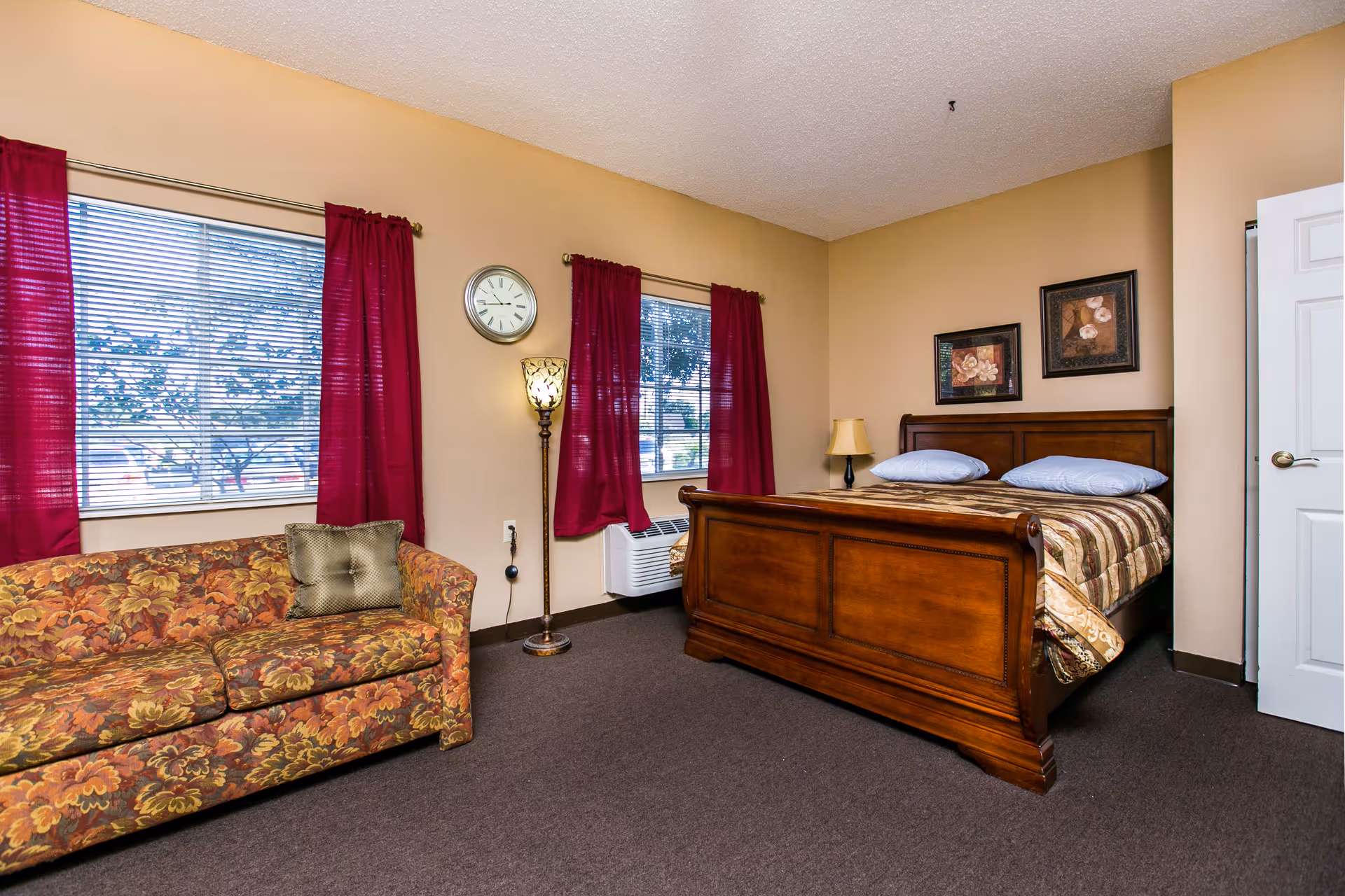 A bedroom with a wooden bed frame and a patterned bedspread, two pillows, two framed floral pictures on the wall above the bed, a floral-patterned couch with a decorative pillow, two windows with red curtains, a floor lamp, a wall clock, and a beige carpeted floor.