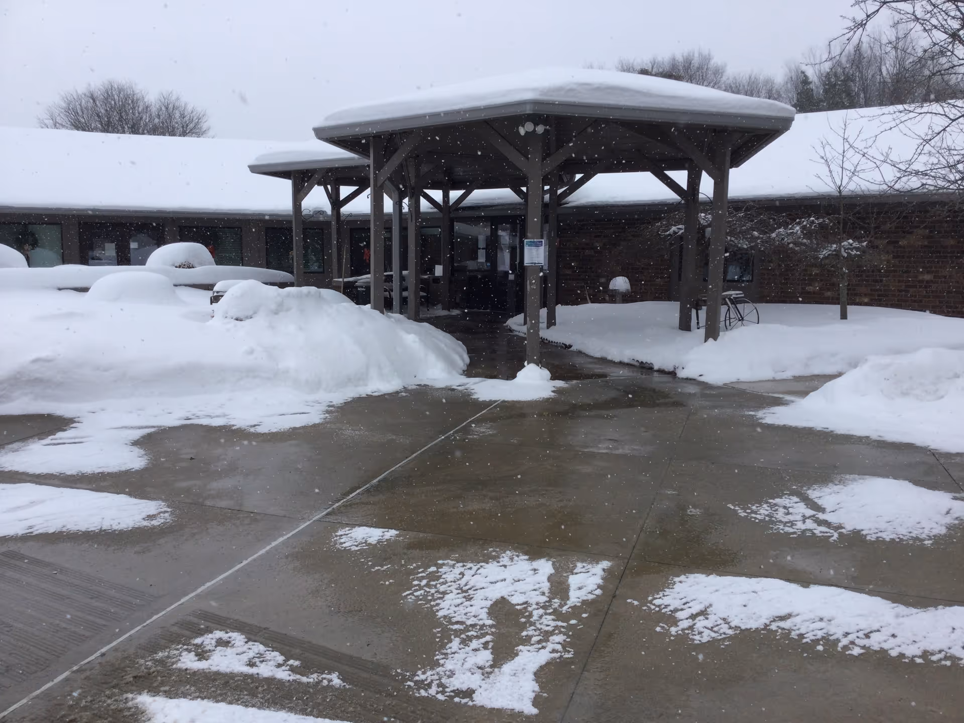 Snow-covered entrance of a single-story brick building with a covered walkway supported by wooden beams. Snow is piled on the ground and roof, and snowflakes are falling. Bare trees are visible in the background.