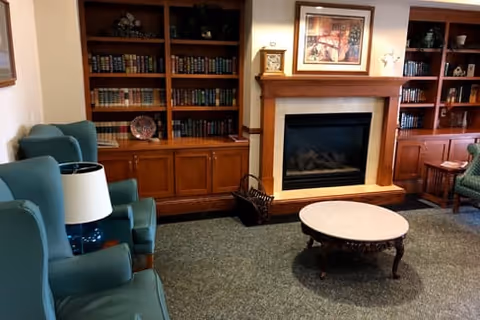 A cozy living room area with green upholstered armchairs, a round wooden coffee table, a fireplace with a wooden mantle, and built-in wooden bookshelves filled with books and decorative items.