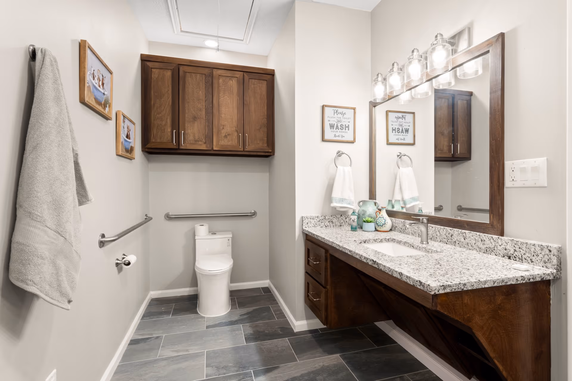 Accessible bathroom featuring a toilet with grab bars, a granite countertop vanity with large mirror and wood cabinets.