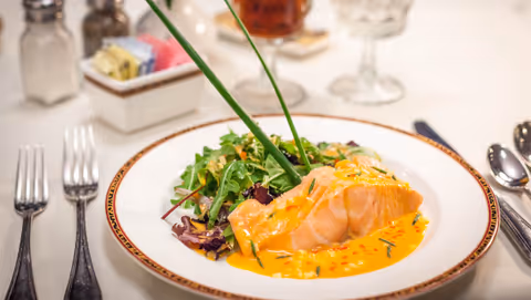 A plated dish featuring a piece of cooked salmon topped with a creamy orange sauce, served alongside a fresh green salad with mixed greens. The plate is set on a white tablecloth with silverware, salt and pepper shakers, and a glass of iced tea in the background.