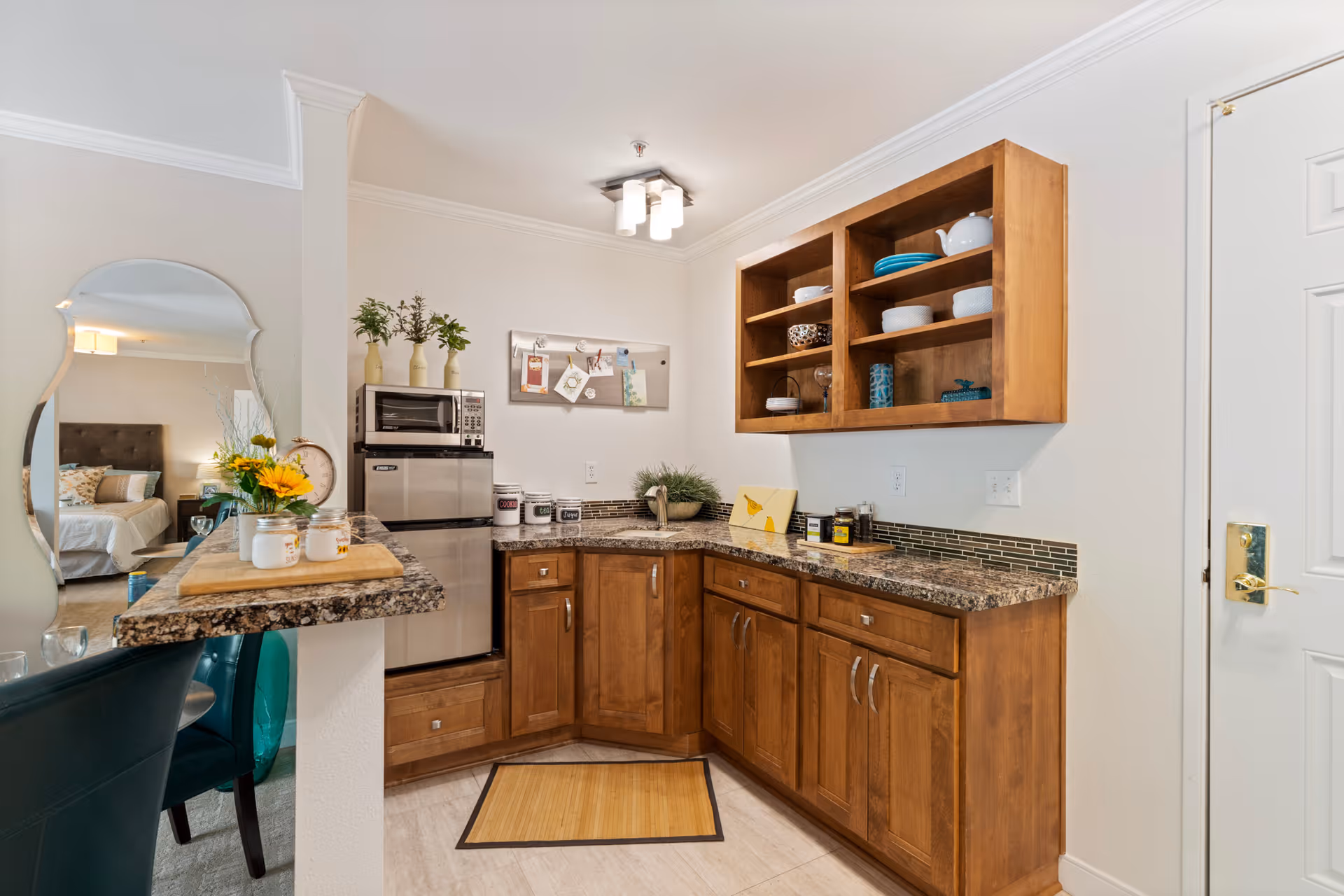 A compact kitchen area with wooden cabinets and granite countertops. There is a small stainless steel refrigerator and microwave stacked on top of each other. Open shelves above the counter hold various dishes and bowls. A small bulletin board with notes is mounted on the wall. To the left, a breakfast bar with a granite countertop has two teal chairs and a vase with yellow flowers. A mirror on the left wall reflects part of a bedroom with a bed and nightstand.