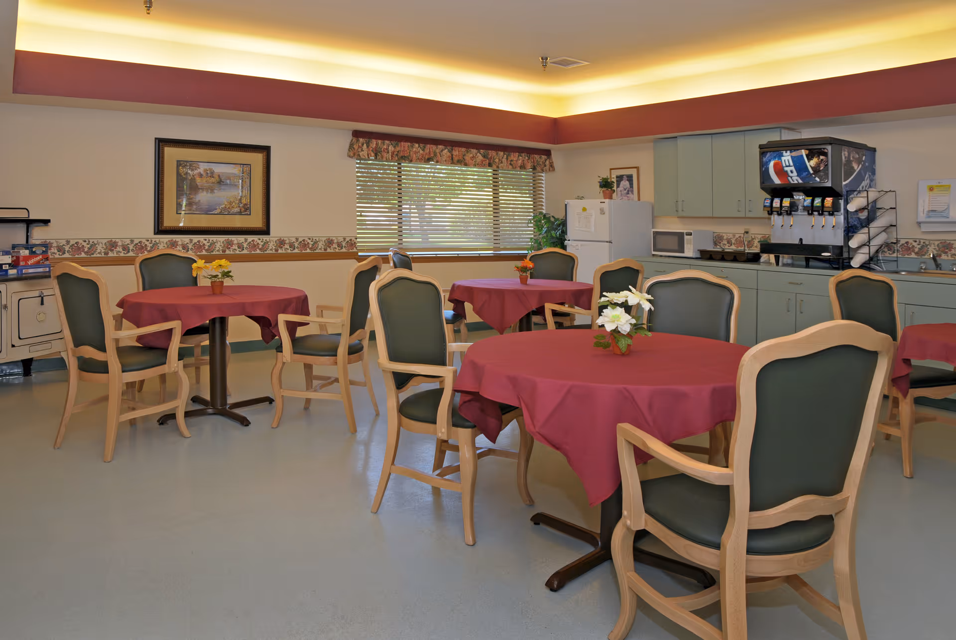 A dining room with several round tables covered with red tablecloths, each table decorated with a small flower arrangement. The room has green cushioned wooden chairs around the tables, a window with blinds and floral valance, a beverage dispenser, a microwave, a refrigerator, and cabinets along one wall. The ceiling has recessed lighting with a warm glow.