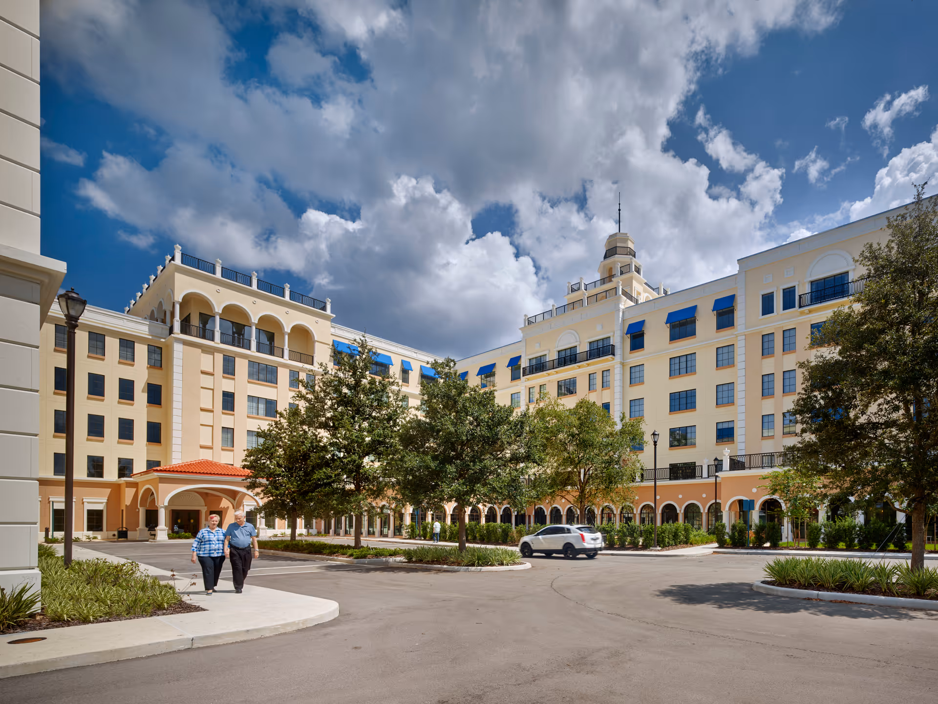 A large multi-story senior living building with an arched entry, landscaped trees, a parked car and two people walking in front under a partly cloudy sky.