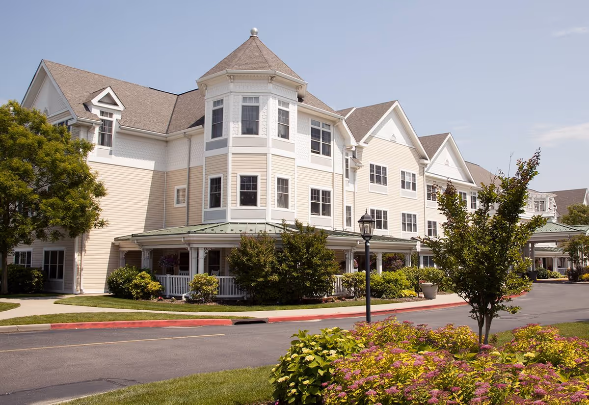Exterior view of a large, multi-story senior living facility with beige siding, white trim, and a turret-like architectural feature. The building is surrounded by well-maintained landscaping including bushes, trees, and flowering plants. A paved driveway and sidewalk are visible in front of the building under a clear sky.