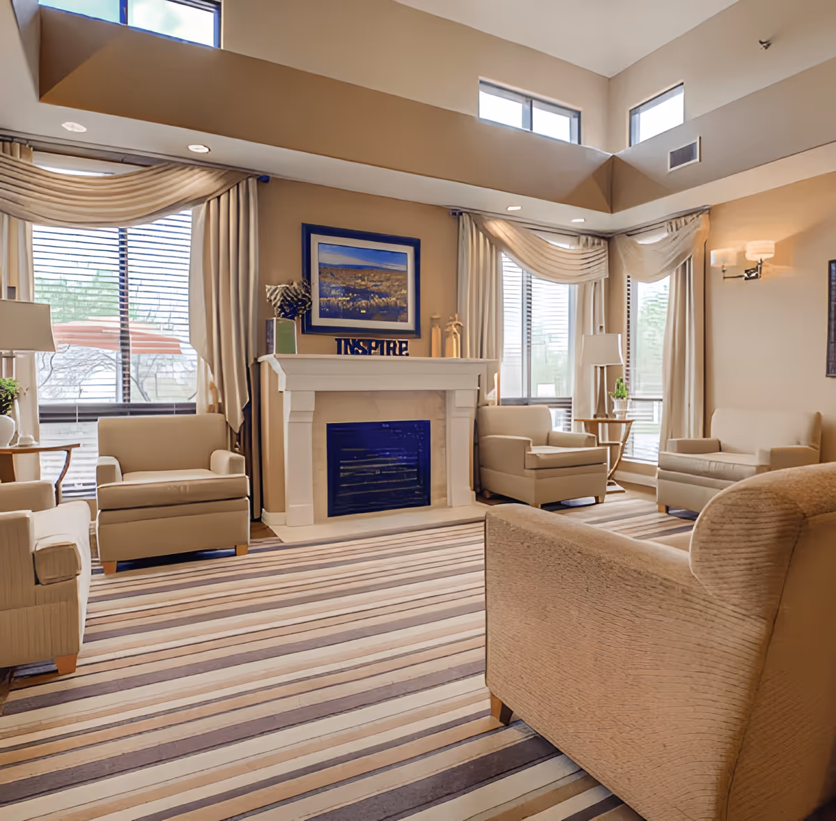 Bright, spacious common room with armchairs arranged around a fireplace, large windows, and a striped carpet.