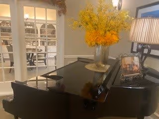 A black grand piano with a vase of yellow flowers on top, a framed photo, and a lamp with a striped shade. In the background, there is a room with white shelves and a desk visible through glass-paneled double doors.