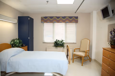 A simple, clean bedroom in a senior living facility with a single bed covered in a light blue quilt, a wooden chair with cushioned seat and backrest, a wooden dresser with a potted plant on top, a blue wardrobe, and a window with blinds and a decorative valance.