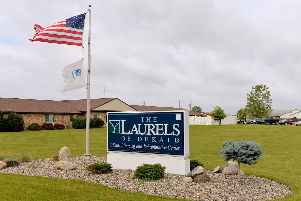 Outdoor view of The Laurels of DeKalb skilled nursing and rehabilitation center sign with an American flag and another flag on a flagpole, surrounded by a landscaped area with rocks and small bushes, with the facility building and parked cars in the background under a cloudy sky.