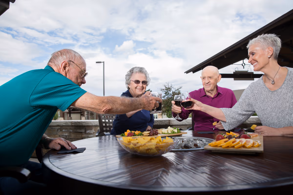 Four elderly people sitting around a round outdoor table, clinking glasses of red wine in a toast. The table has plates of snacks including chips, cheese, grapes, and fruit. The setting is outside with a partly cloudy sky and a building structure visible in the background.