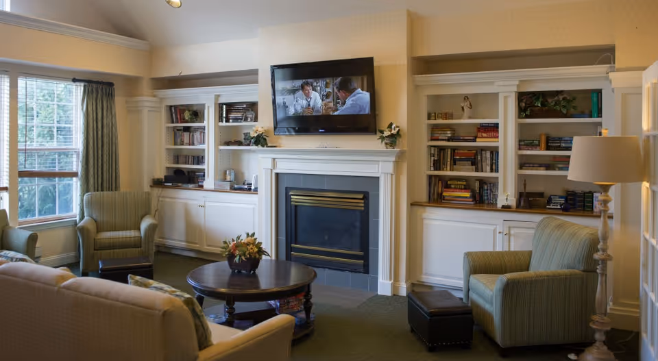 Cozy communal living room with armchairs and sofas arranged around a fireplace with a wall-mounted TV, bookshelves, a coffee table, and a large window.