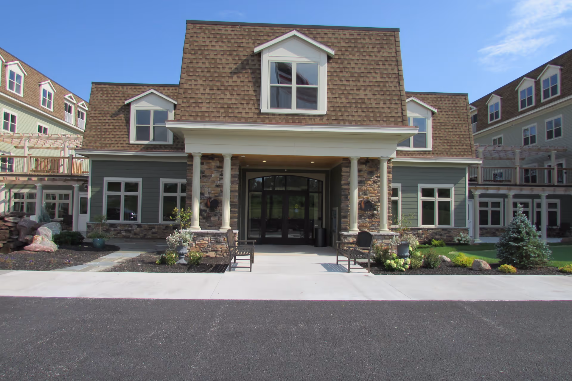 The front entrance of a multi-story residential building with a covered portico, columns, benches, and landscaped beds.