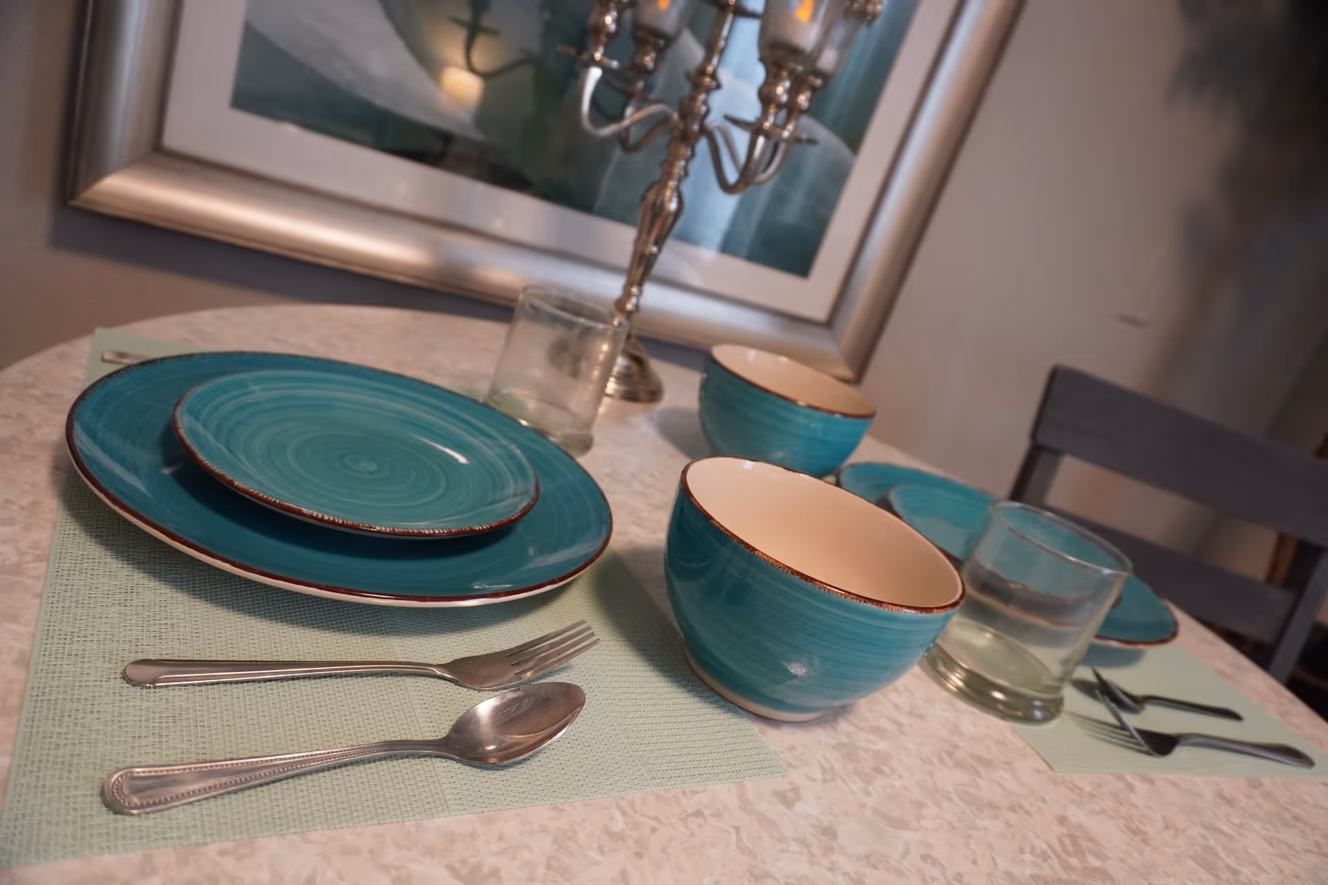 A dining table set for two with turquoise plates and bowls, silver fork and spoon, clear drinking glasses, and a silver candelabra. The table has light green placemats and is positioned in front of a framed artwork on the wall.