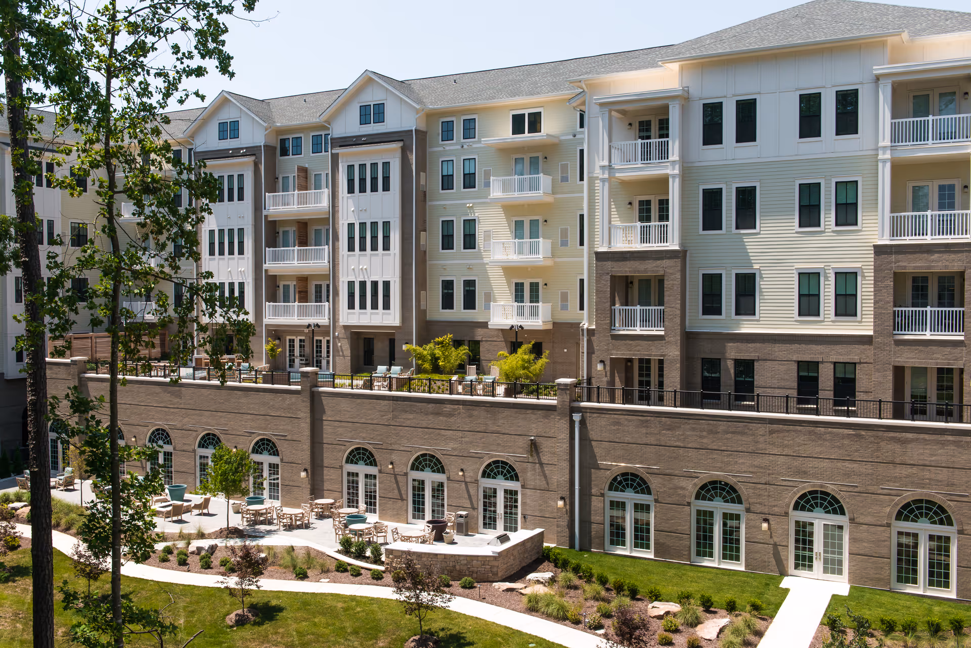 Exterior view of a multi-story senior living facility with balconies and large arched windows on the lower level. The building is surrounded by landscaped gardens with walking paths, outdoor seating areas, and trees under a clear sky.