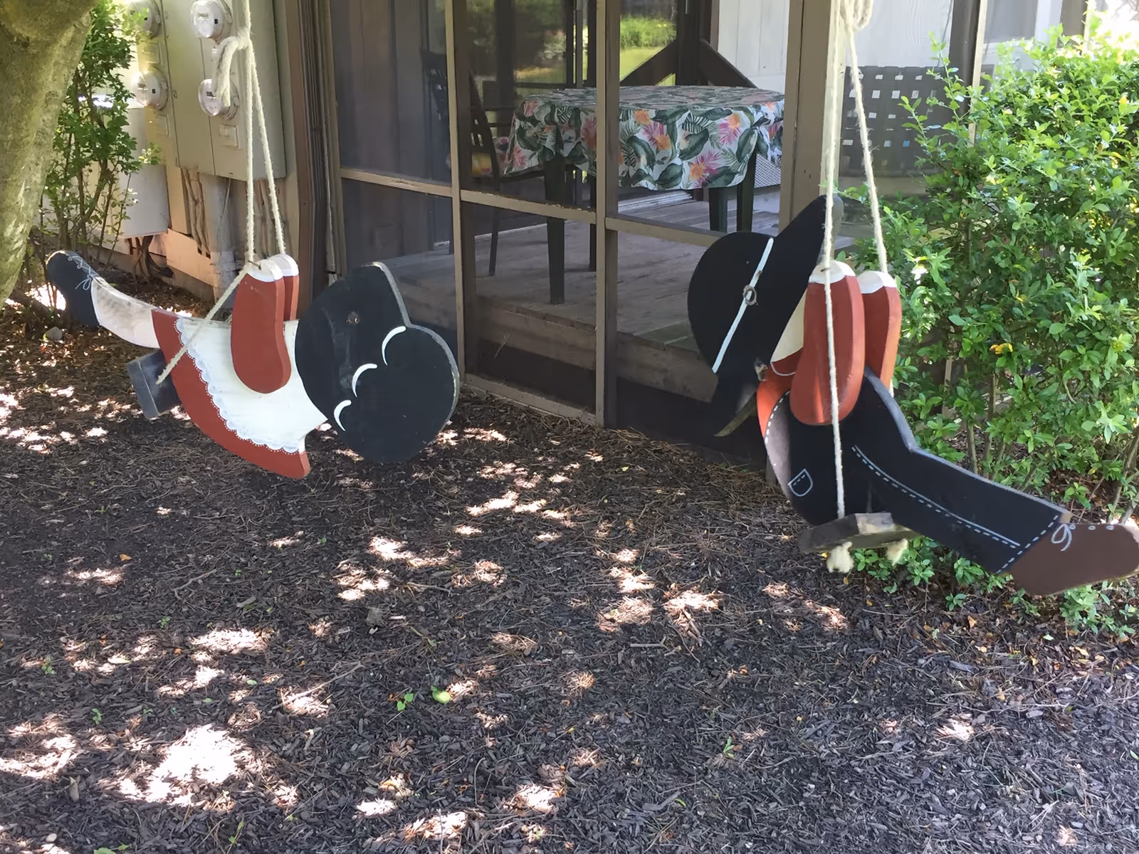 Two decorative wooden cutout swings shaped like children hang from ropes under a tree in front of a screened porch.