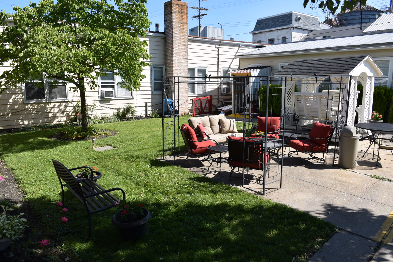 Outdoor patio area with green grass, a tree, and various seating options including a black metal bench, cushioned chairs with red pillows, a beige cushioned loveseat, and a white wooden swing under a small pergola. The area is adjacent to a light-colored building with windows and a chimney.