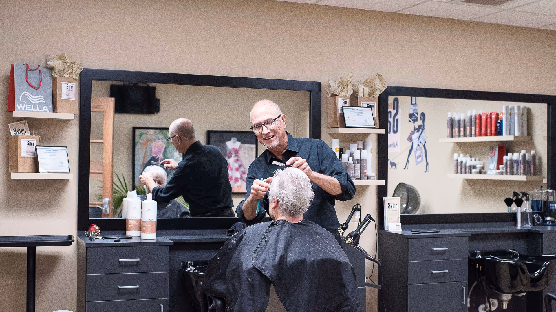 A hairstylist cutting the hair of an elderly man in a salon area with large mirrors, shelves holding hair care products, and a black hair washing sink.