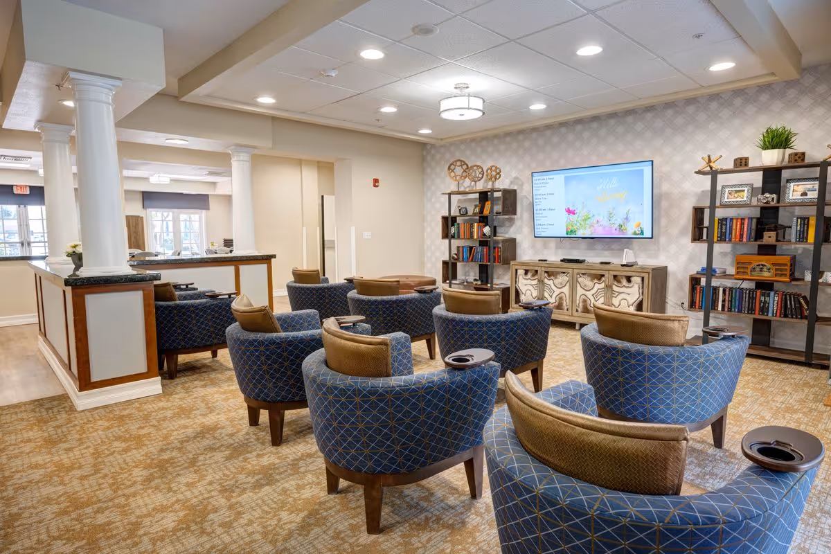 A cozy common area in a senior living facility featuring several blue upholstered armchairs arranged in rows facing a wall-mounted flat screen TV. The room has beige patterned carpet, light-colored walls with subtle wallpaper, and two tall bookshelves filled with books and decorative items. There are white columns and a partial view of another seating area in the background.