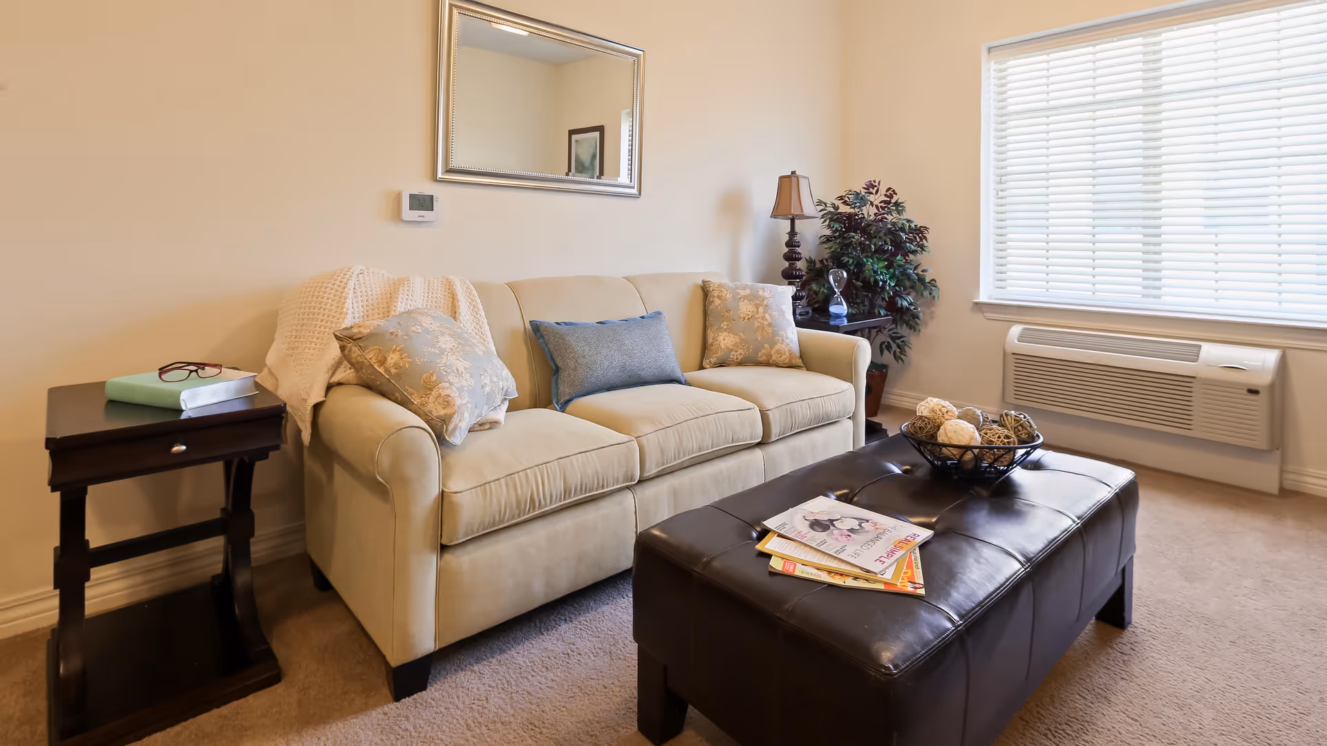 A cozy living room with a beige sofa adorned with decorative pillows and a throw blanket. Next to the sofa is a dark wooden side table with a book and glasses on it. In front of the sofa is a dark brown leather ottoman with magazines and a decorative bowl on top. A window with blinds and a wall-mounted air conditioning unit are visible on the right side. A potted plant and a table lamp are placed in the corner near the sofa.
