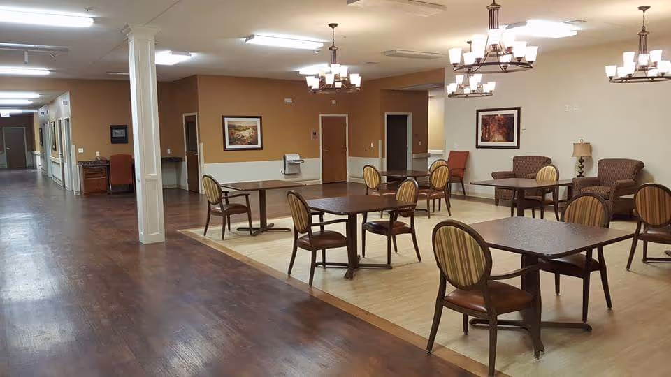 Interior view of a nursing and rehab facility common area with several tables and chairs arranged on a light wood floor section. The surrounding floor is darker wood. There are chandeliers hanging from the ceiling, framed artwork on the walls, and a hallway leading to other rooms.