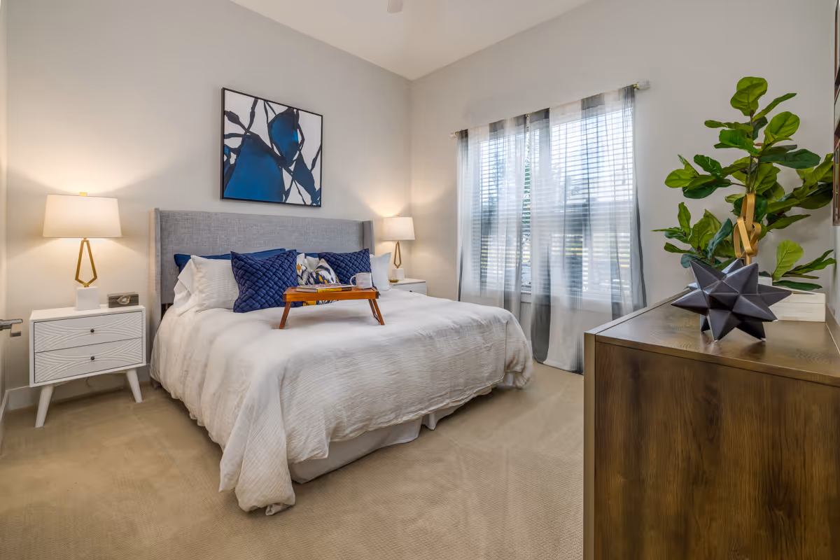 Well-lit bedroom with a made bed, bedside tables and lamps, artwork over the headboard, a window with sheer curtains, and a dresser with a plant.