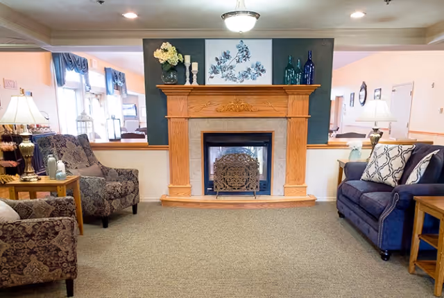 A cozy living room area with a wooden fireplace mantle decorated with vases and floral artwork above it. There are two patterned armchairs on the left and a blue sofa with patterned pillows on the right. Side tables with lamps and decorative items flank the seating. The room has a carpeted floor and soft lighting.