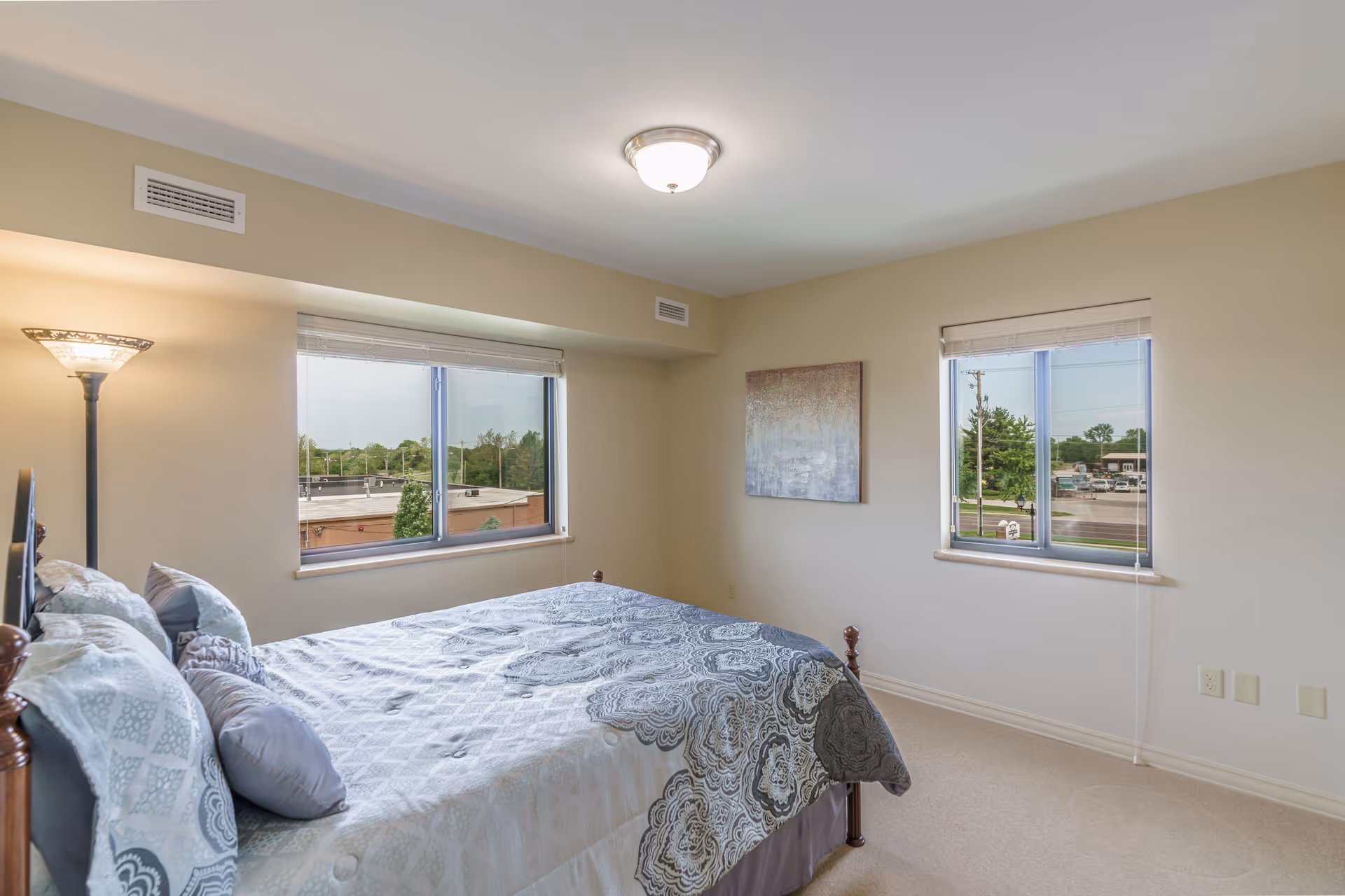 A bedroom with a bed covered in a patterned gray and white bedspread, two windows with views of trees and buildings outside, a floor lamp in the corner, and a piece of abstract artwork on the wall.