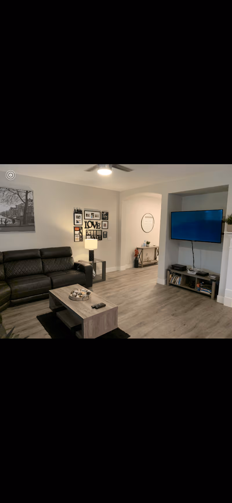 A modern living room with a black leather sofa, a wooden coffee table with decorative items, a wall-mounted flat-screen TV, and a small entertainment unit with books and devices. The room has light-colored wooden flooring, a ceiling fan with light, and wall decorations including framed photos and a large black and white picture.