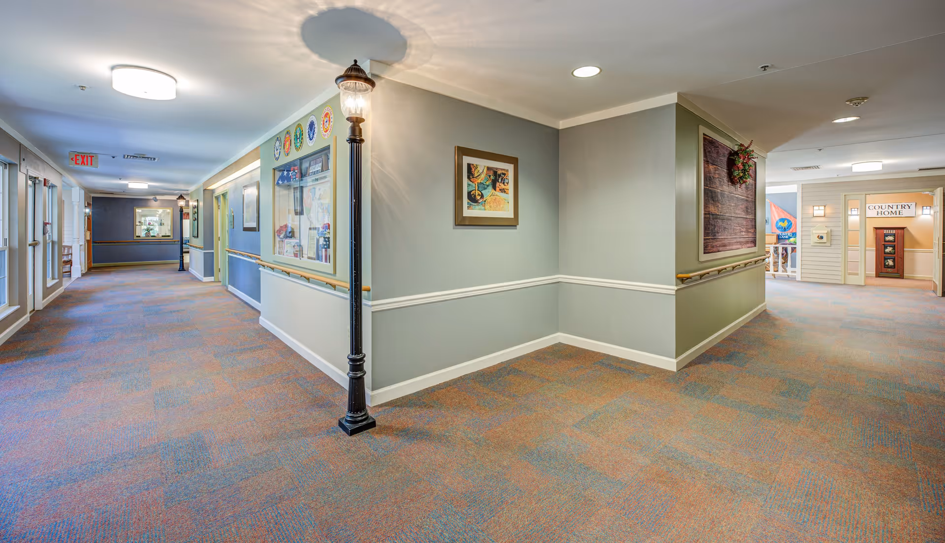 A well-lit interior hallway in a senior living facility with light blue walls, handrails, and colorful carpet. The hallway features framed artwork, a decorative streetlamp-style light fixture, and various doors and windows along the walls. An exit sign is visible at the far end, and a sign reading 'Country Home' is seen in the distance.