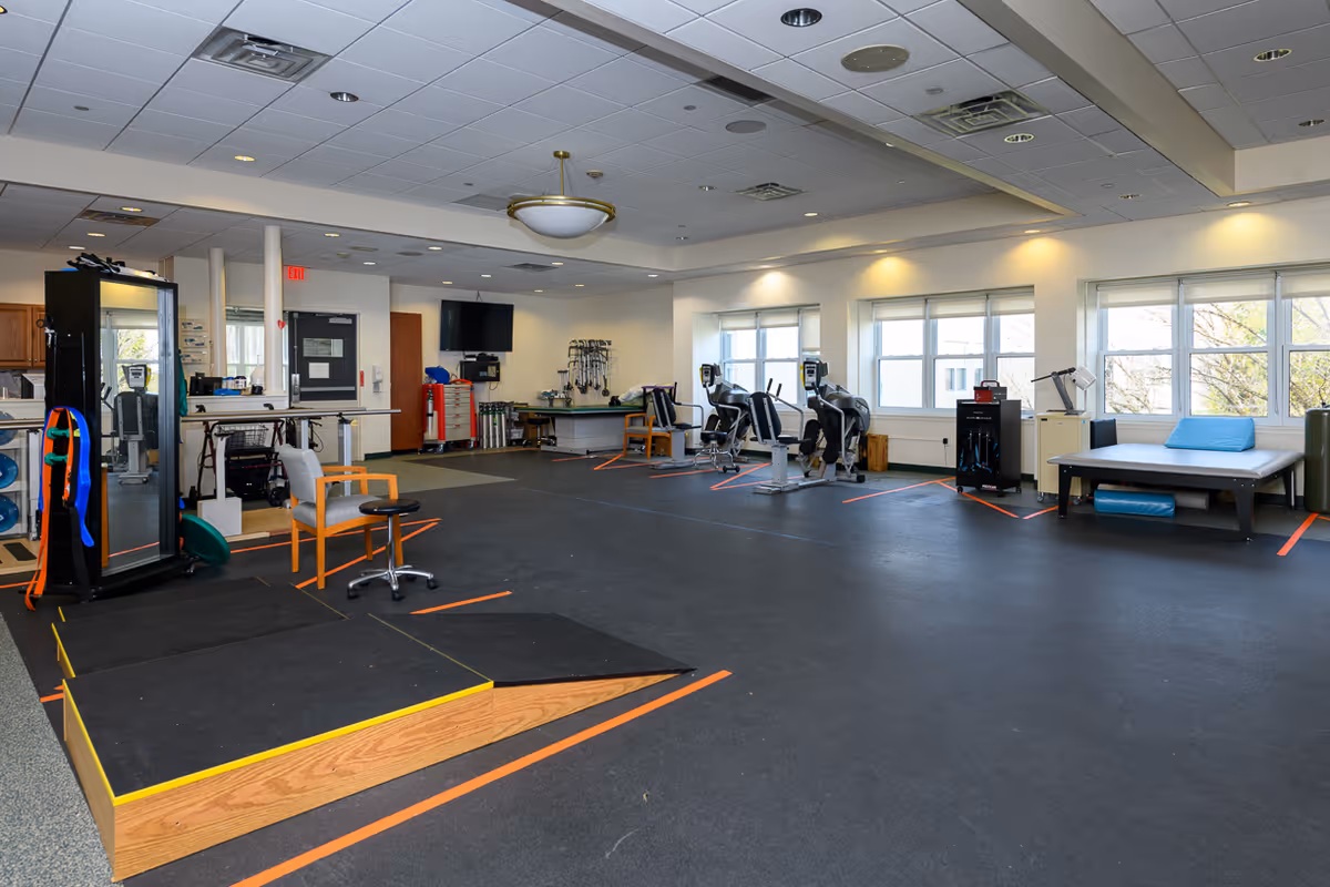 A spacious rehabilitation and physical therapy room with exercise equipment including stationary bikes, a therapy bed with blue cushions, chairs, and a small raised platform with ramps. The room has large windows letting in natural light and a TV mounted on the wall.