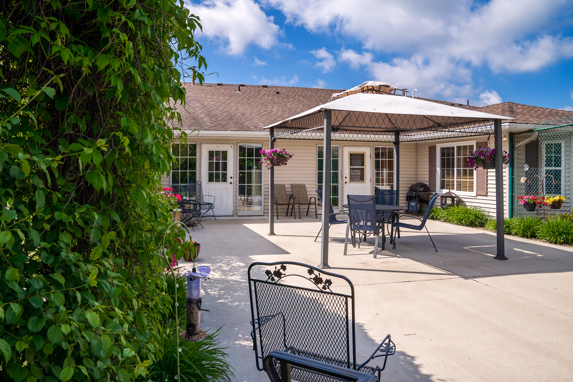 Outdoor patio area at The Glenn Buffalo with a metal gazebo covering a table and chairs. There are hanging flower pots, a barbecue grill, and additional seating along the building's exterior. The patio is surrounded by greenery and plants, with a clear blue sky overhead.