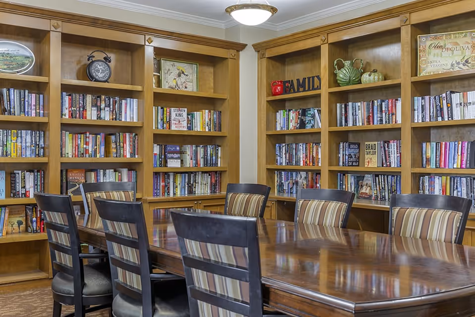 A cozy library room with wooden bookshelves filled with books and decorative items. In front of the shelves is a polished wooden table surrounded by six chairs with striped cushions. The room has a warm and inviting atmosphere with soft lighting from a ceiling fixture.