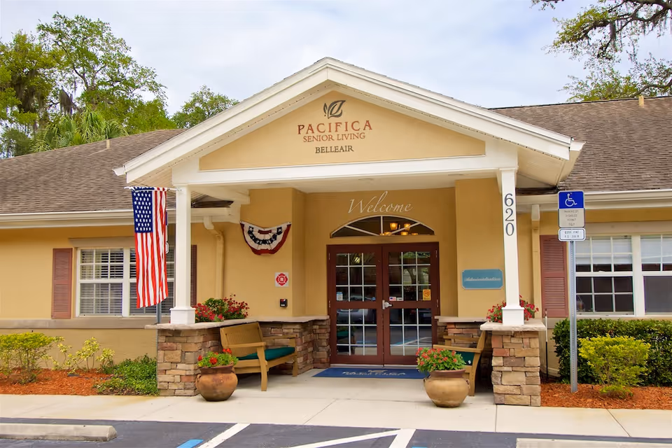 Entrance of Pacifica Senior Living Belleair building with a covered porch, two benches, potted plants, an American flag, and a handicap parking sign. The building is beige with stone accents and has the number 620 on a pillar.