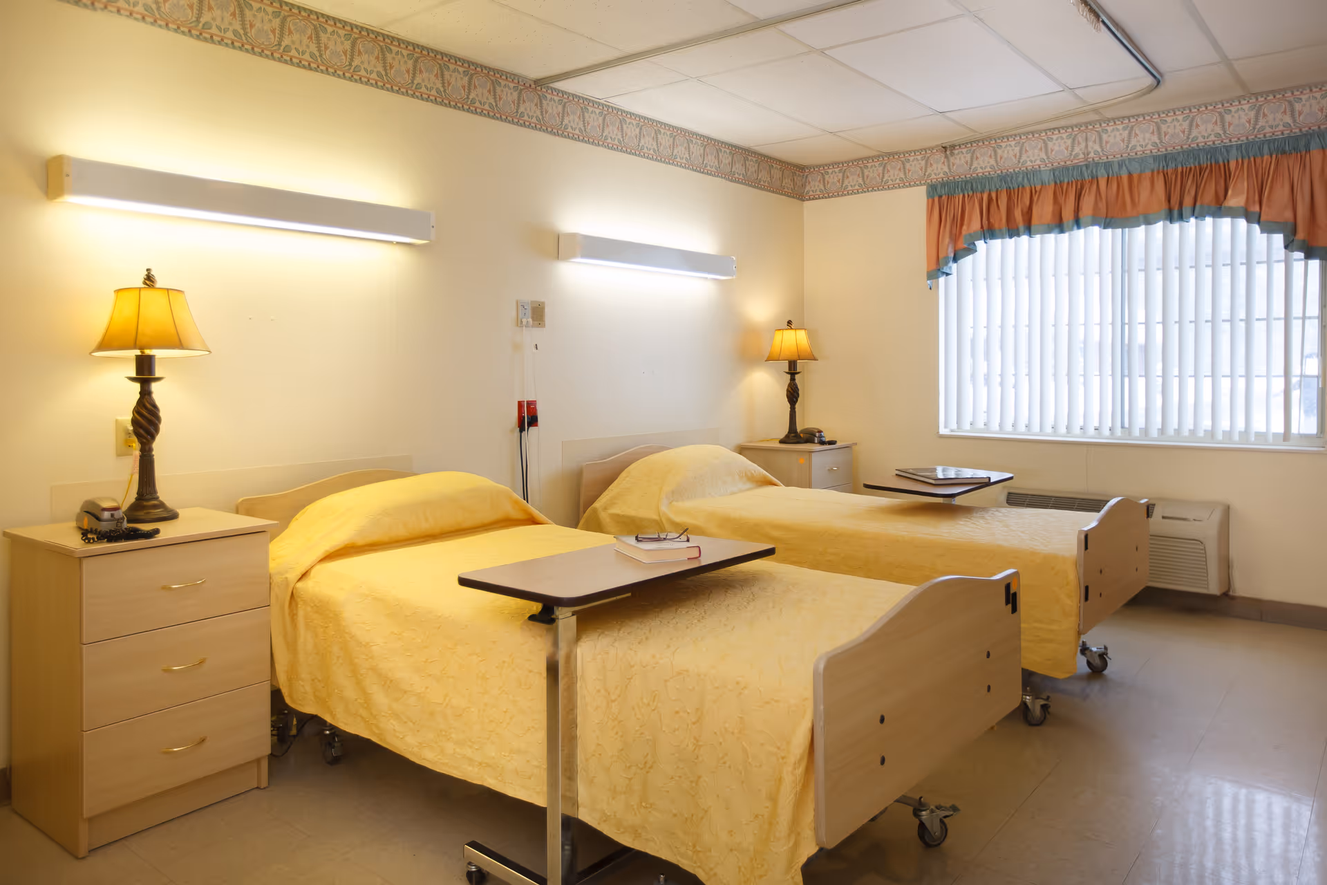 A bright room with two single beds covered in yellow bedspreads, each with an overbed table. There are two bedside tables with lamps, a window with vertical blinds and a valance, and fluorescent lights on the wall above the beds.