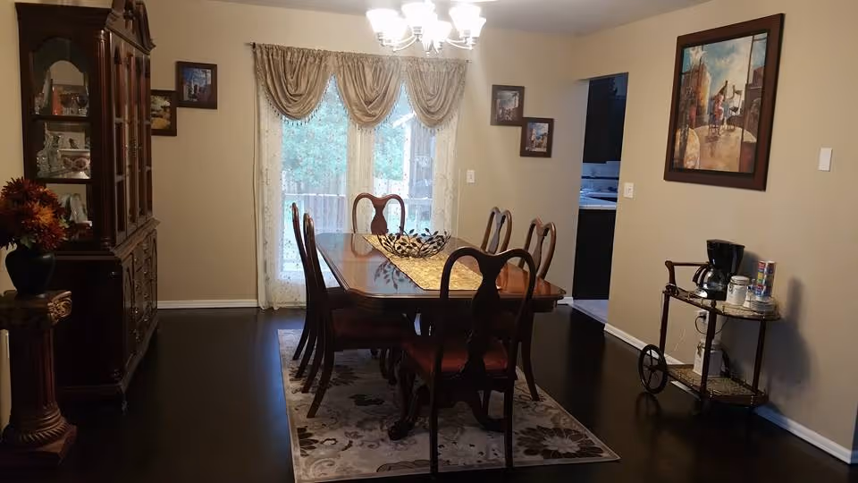 A dining room with a wooden dining table and six chairs placed on a patterned rug. The room has beige walls with framed artwork and a large window with sheer curtains and valances. There is a wooden china cabinet on the left side and a small serving cart with a coffee maker and supplies on the right side. A chandelier hangs from the ceiling providing light.