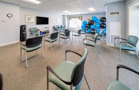 Bright multipurpose activity room with spaced green chairs arranged in a circle, a wall-mounted TV, exercise balls and a water cooler.