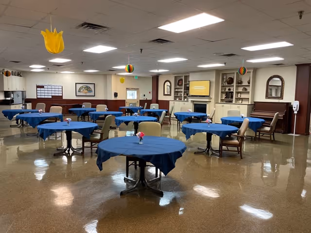 A spacious dining room with multiple round tables covered in blue tablecloths, each with a small flower centerpiece. The room has beige walls with dark wood paneling, a piano, a fireplace with a TV above it, and decorative mirrors on the walls. Colorful hanging decorations are suspended from the ceiling.