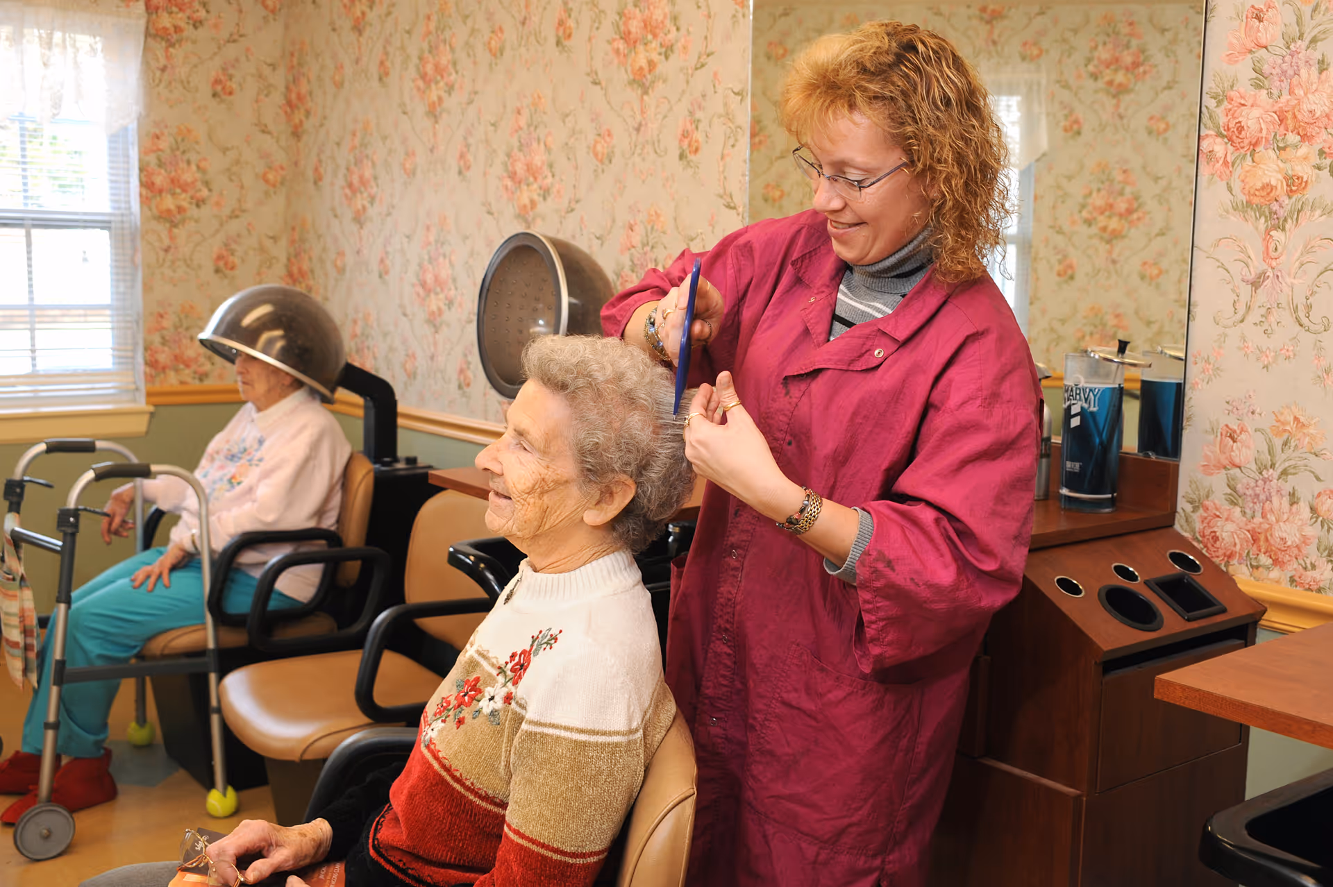 A hairdresser in a magenta smock is styling the hair of an elderly woman seated in a chair, smiling. Another elderly woman is sitting under a hair dryer in the background. The room has floral wallpaper and a window with blinds.