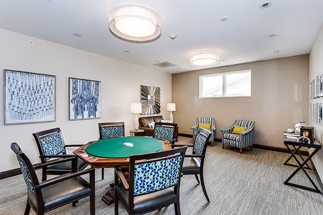 Well-lit common room with a round green card table surrounded by patterned chairs and additional armchairs and lamps.