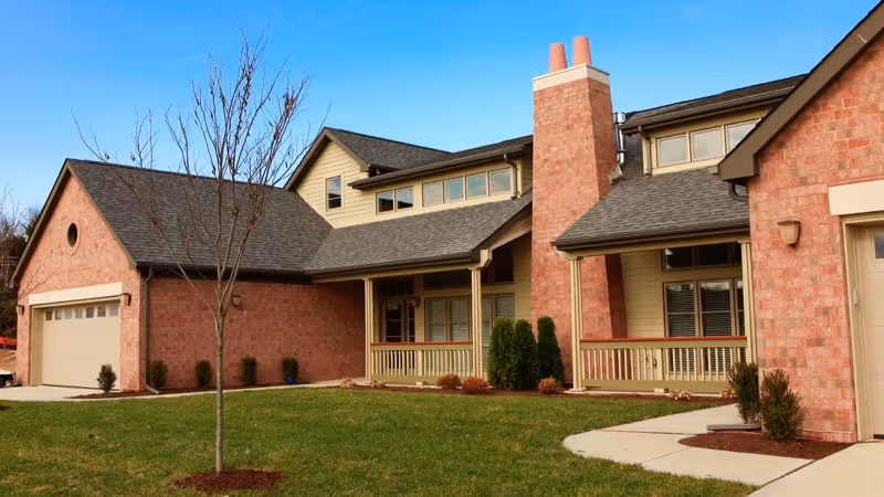 Exterior view of a residential-style building with brick and beige siding, featuring a chimney, garage, and a small porch area. The building is surrounded by a well-maintained lawn with a young tree and some shrubs under a clear blue sky.