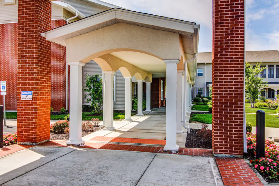 Covered entrance walkway with white columns at the front of a red-brick senior living building and landscaped grounds.