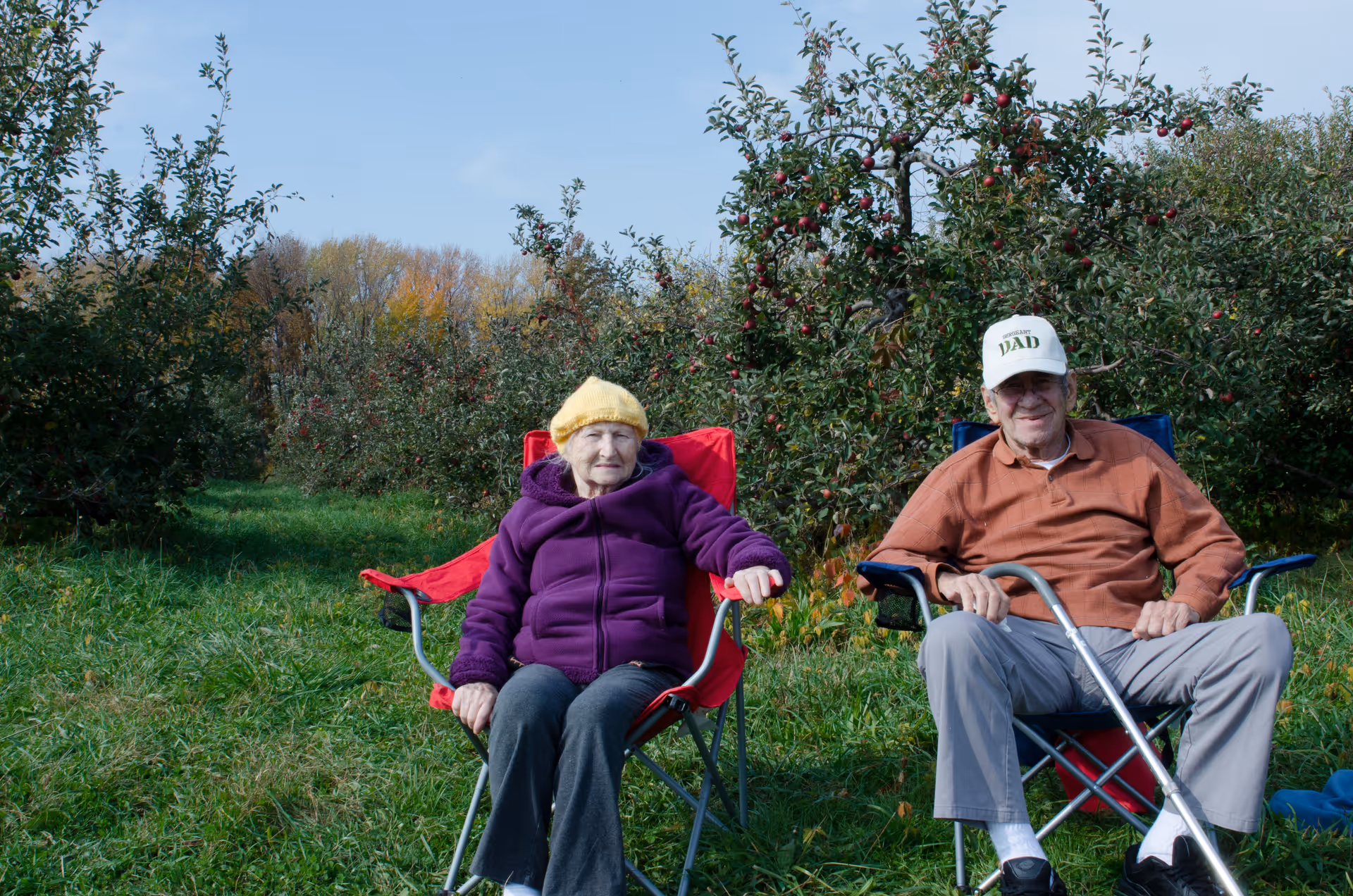 Two elderly individuals sitting outdoors on foldable chairs in an apple orchard. The woman on the left is wearing a yellow knit hat and a purple jacket, while the man on the right is wearing a white cap, orange shirt, and holding a cane. Apple trees with red apples are visible in the background under a clear blue sky.