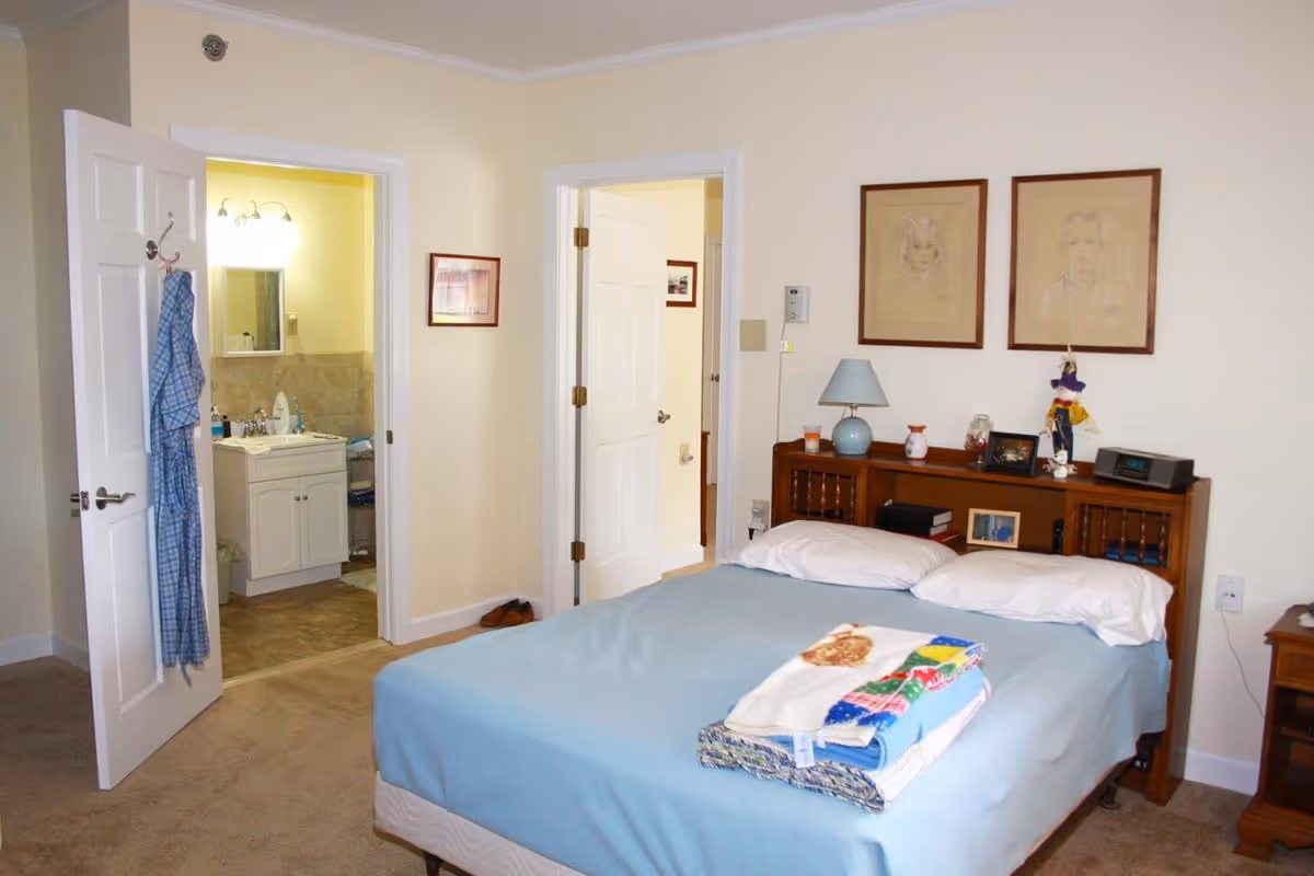 A furnished bedroom with a made bed, headboard and bedside table, with doors open to a bathroom and a hallway.