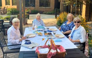 Four elderly women sitting around a rectangular outdoor dining table covered with a white tablecloth, set with plates and glasses, enjoying a sunny day at an outdoor patio area with brick buildings and greenery in the background.