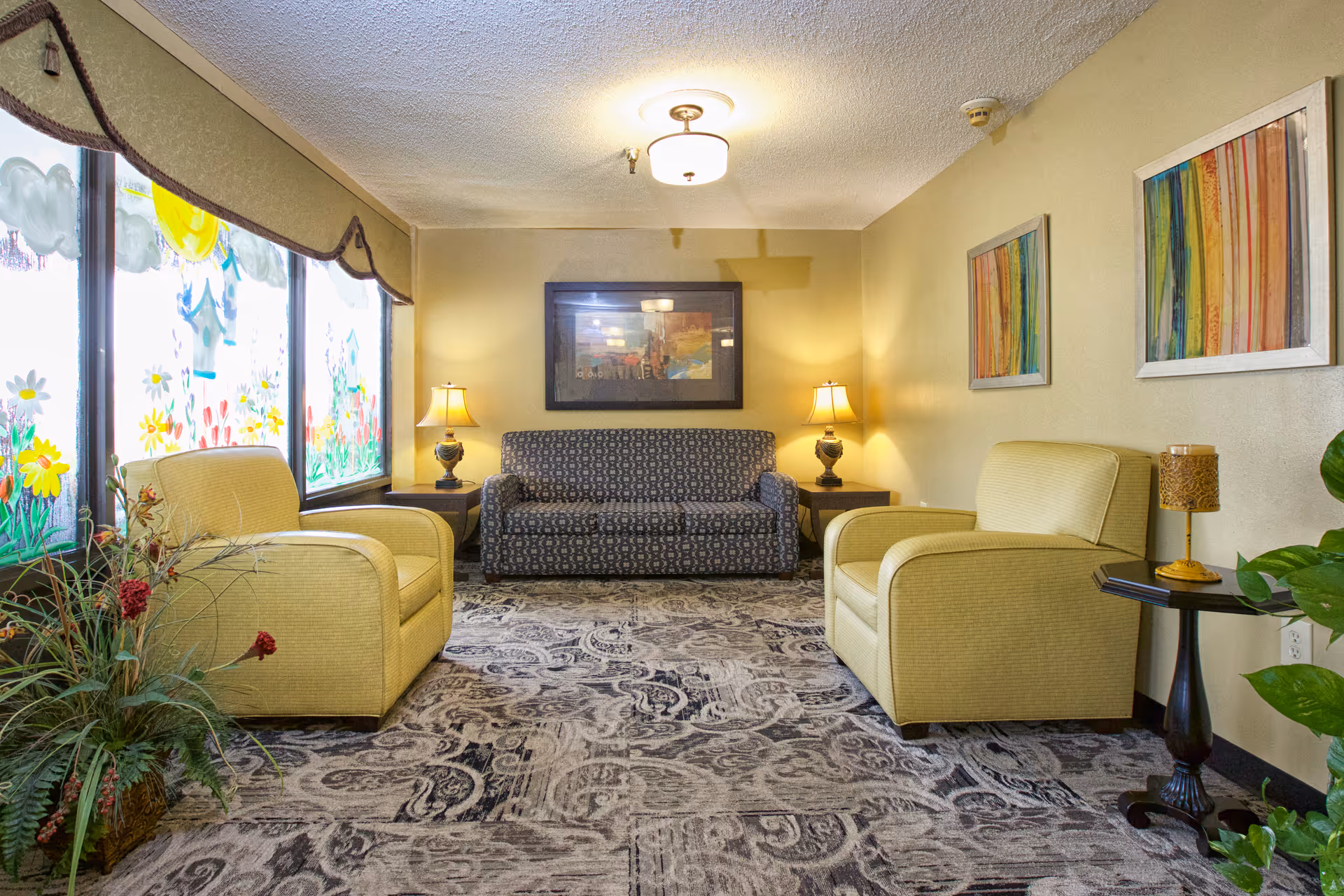 A cozy living room area with two yellow armchairs facing a patterned dark sofa. The room has a patterned carpet, two side tables with lamps, colorful abstract paintings on the walls, and large windows decorated with cheerful flower and sun artwork. There are also some green plants adding a touch of nature to the space.